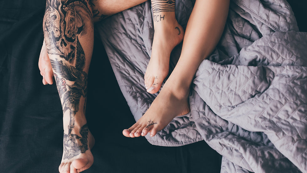 Dark purple and lilac bedsheets with the lower legs of a couple with tattoos are visible.