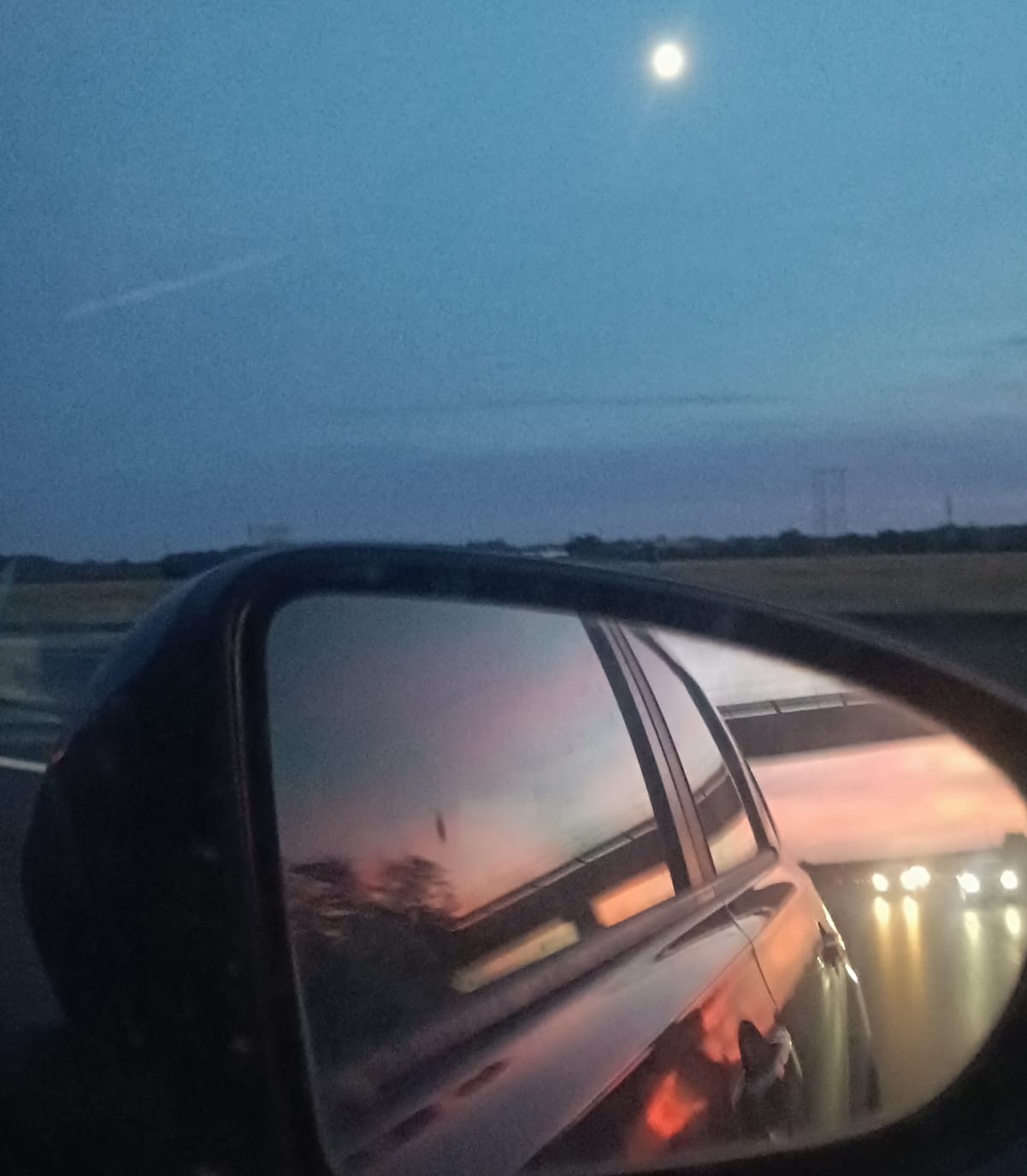 Route au crépuscule vue depuis une voiture, avec la lune dans le ciel et le reflet de la voiture dans le rétroviseur.