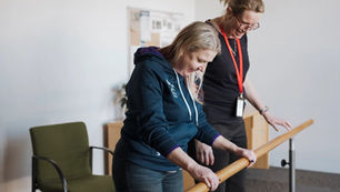 A support worker helps a middle aged woman to walk, both hanging on to a hand rail