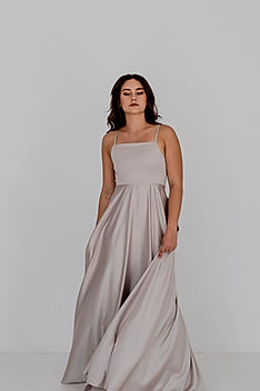 Model wearing the Gelique Esme Dress in a soft taupe satin, featuring a minimalist square neckline, thin straps, fitted bodice, and a flowing full-length skirt with elegant drape. Photographed against a clean white studio background.