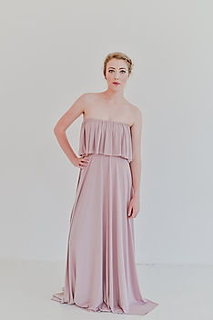 Model wearing the Gelique Jessy Dress in a soft dusty mauve shade. The gown features a strapless neckline with a flowing ruffle overlay that can be styled on one shoulder, both shoulders, or off-the-shoulder for versatile bohemian elegance. Underneath the frill is a fitted bodice that smooths the waist and transitions into a long, fluid A-line skirt that drapes beautifully to the floor. Photographed against a clean white studio backdrop to emphasize the dress’s soft movement and romantic silhouette.