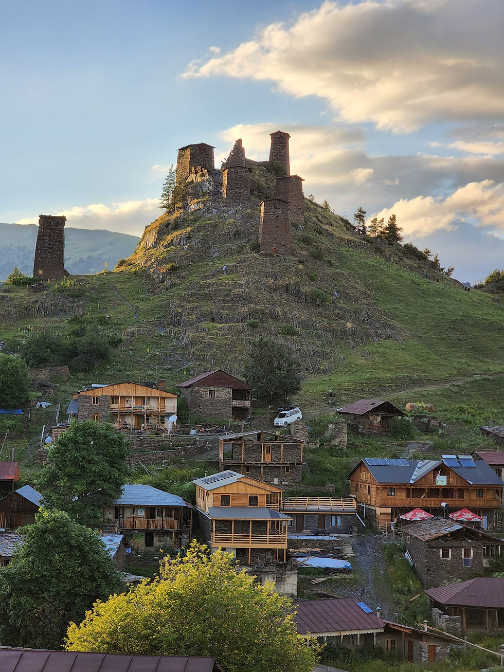 Stone towers on a green hill overlook a village of wooden houses under a blue sky with clouds, creating a serene and historic scene.