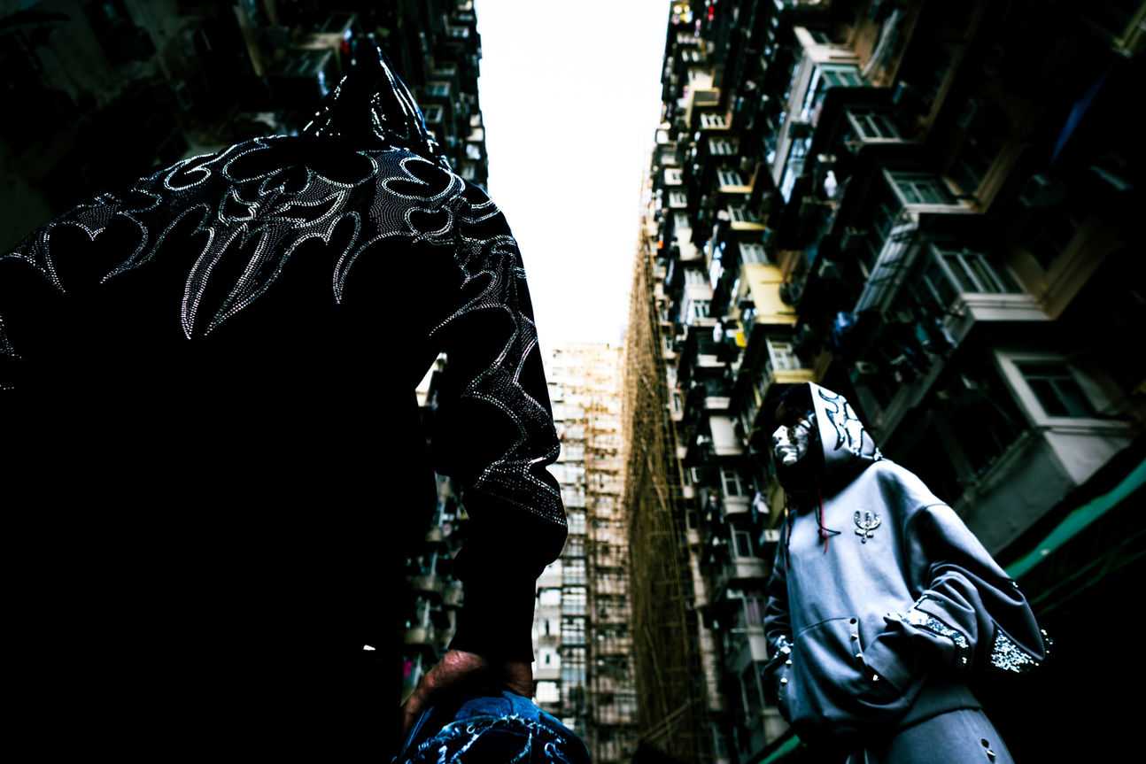 Low-angle shot looking up at the sky from the bottom of Hong Kong's Monster Building (Yick Cheong Building). A model stands in the foreground wearing a black streetwear hoodie featuring a large, sparkling rhinestone tribal skull design on the back. The image captures the dense, claustrophobic urban architecture contrasting with the luxury fashion detail.