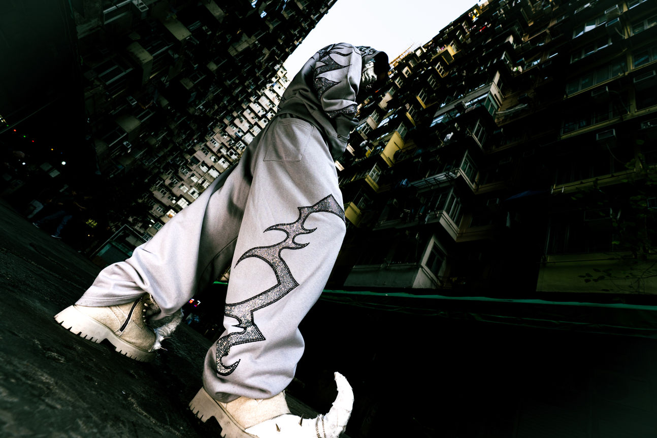 Low-angle detail shot focusing on the leg of a grey streetwear sweatpant. The pant features a large, sharp tribal pattern made of silver rhinestones. The model wears chunky beige boots. The background is the dark, imposing facade of a Hong Kong high-rise building, highlighting the texture and quality of the clothing material.