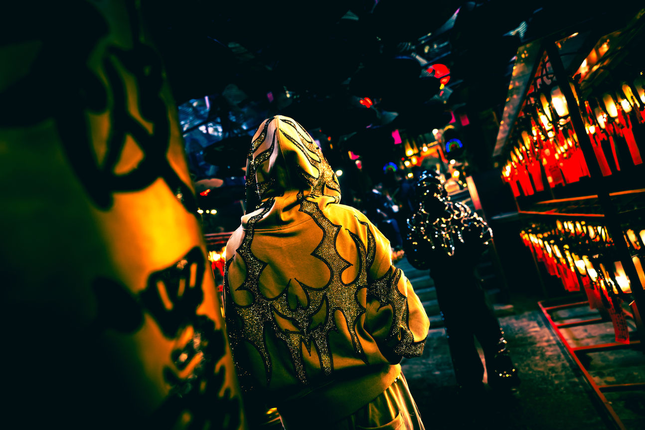 Rear view of a designer hoodie featuring intricate silver stud details, set against a background of blurred red temple lights and dark architecture. High-contrast fashion photography showcasing the texture and quality of the fabric.