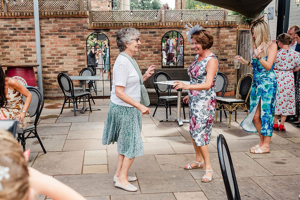 Two women in floral dresses dance joyfully on a patio with brick walls. Other guests chat nearby, creating a lively outdoor gathering.