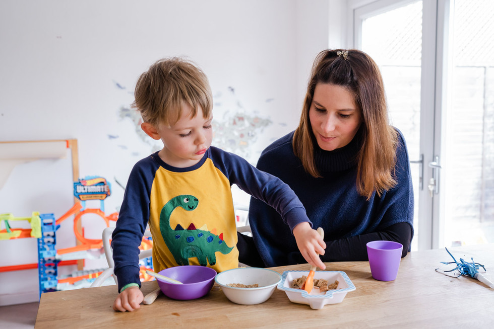 Boy in yellow and blue dinosaur pyjamas miles cheekily at the camera while sitting at a breakfast bar digging a spoon into one of 3 cereal bowls, while his mum sits next to him, watching