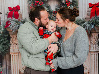 Couple kissing a baby dressed in red in a festive setting with greenery and red bows, creating a warm and joyful holiday mood.