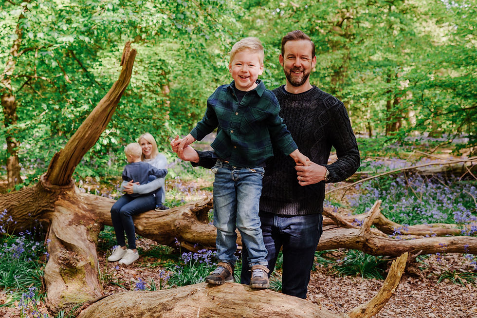 Father and son smiling on a fallen tree in a lush forest. Mother sits with another child in the background. Bright, sunny day.