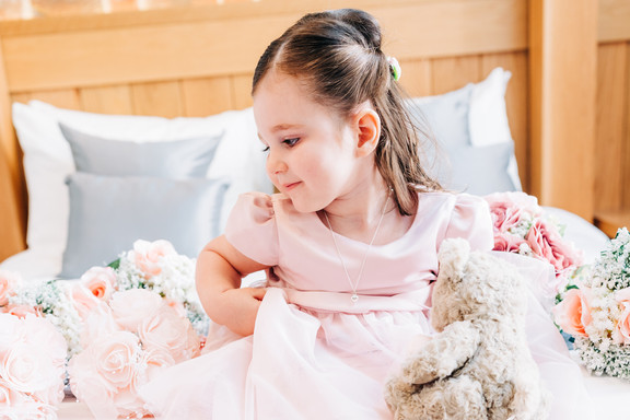 Young flower girl in pink dress sitting on a bed surrounded by wedding flowers