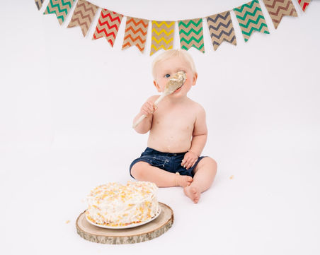 Boy sitting behind a cake in white studio decorated with colourful bunting eating cake fro