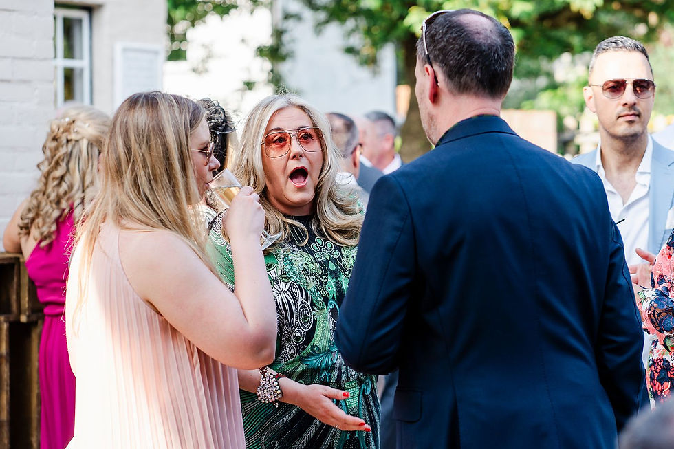 Woman in patterned dress reacts with surprise in a group. Another woman holds a drink. Bright outdoor setting, casual attire, trees visible.