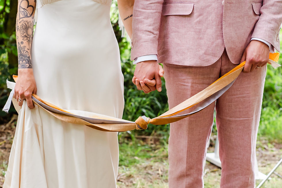 Bride in ivory dress and groom in pink suit hold hands and are linked by natural coloured knotted ribbons. Close up showing hands only. Lush green forest setting.