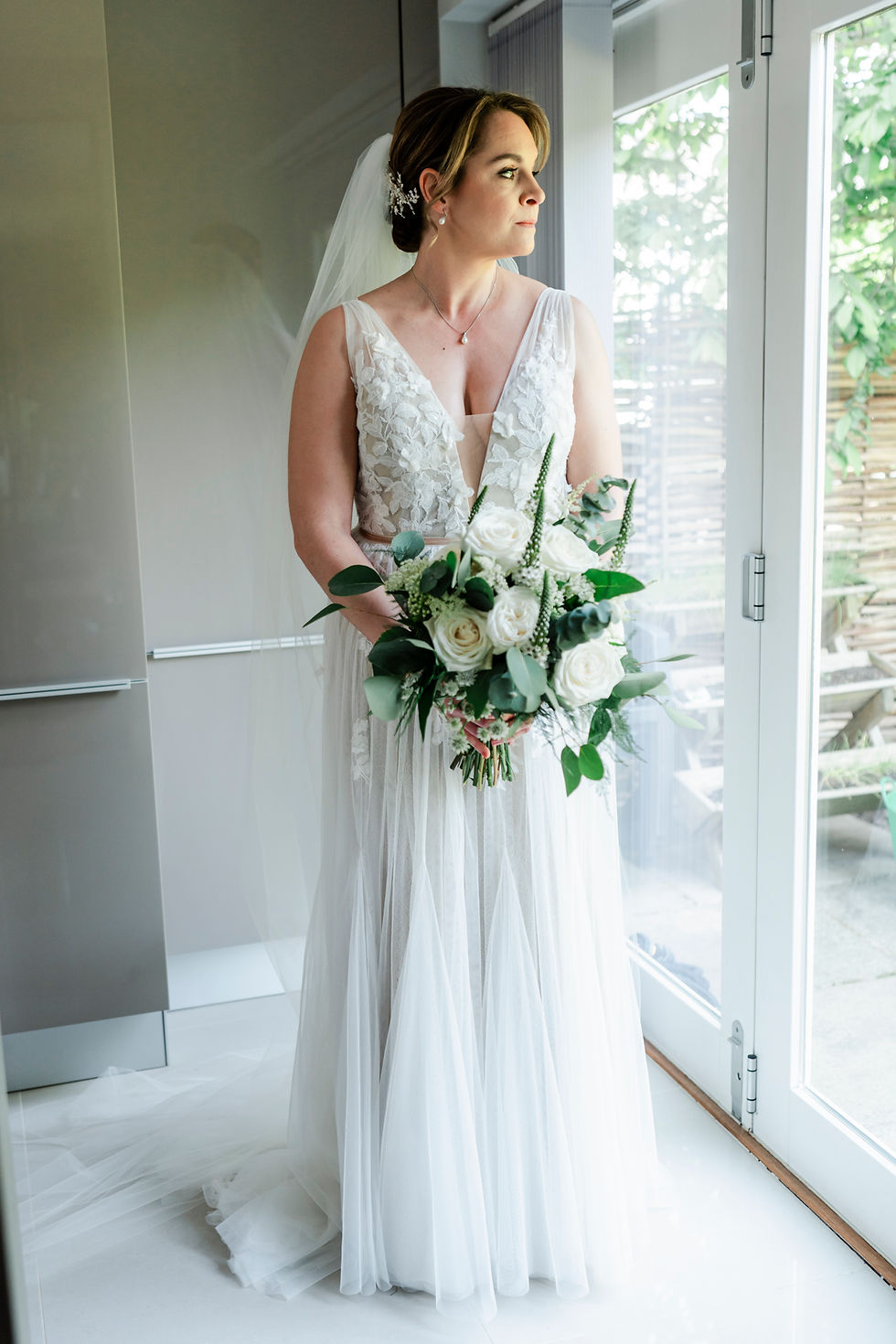 Bride in white dress holding bouquet, looking out glass doors. Light filters in, illuminating her face. Peaceful and reflective mood.
