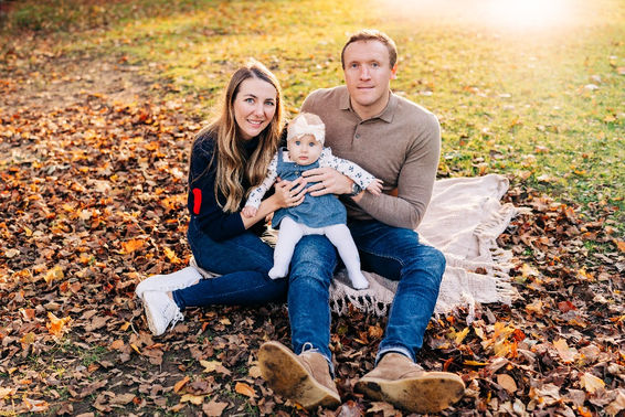 A lady, man and baby girl wearing a pink beanie hat sitting on a blanket outdoors on a bed of autumn leaves
