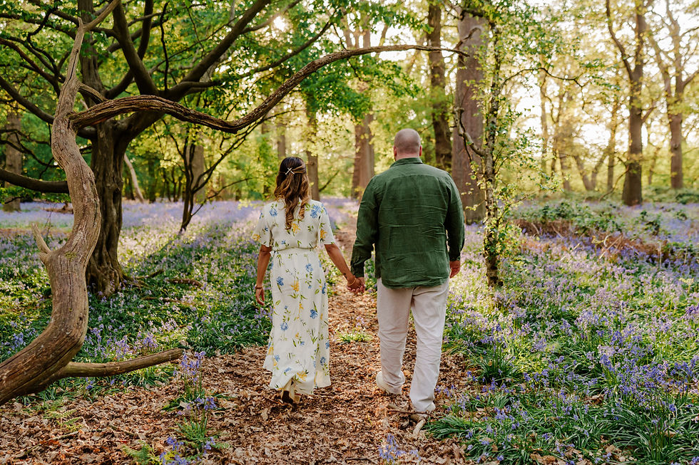 Couple walking hand in hand through a sunlit forest with purple flowers, wearing green and floral outfits. Peaceful, romantic setting.