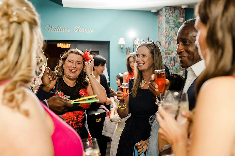 People smiling and holding drinks gather in a room with teal walls and floral decor. The sign reads "Wallace Room." Festive mood.