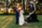 Bride and groom holding hands facing each other with the setting sun peeking through the trees between them
