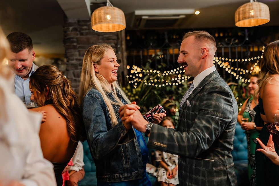 A man and woman joyfully dancing in a warmly lit room with string lights. Others socializing in the background, creating a festive mood.