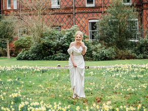 Woman in a white dress stands in a field of yellow daffodils, laughing. A historic brick building is in the background.
