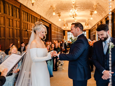 Bride in white gown exchanging rings with groom in navy suit, in an ornate hall. Guests in glittering attire watch, creating a joyful scene.
