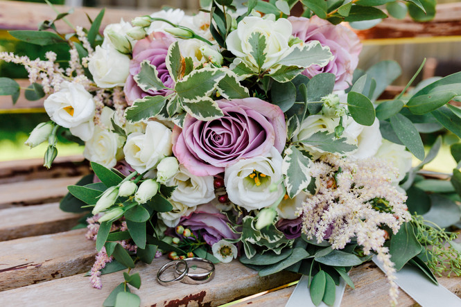 Bouquet of purple roses and white flowers on a wooden bench with two wedding rings in the foreground
