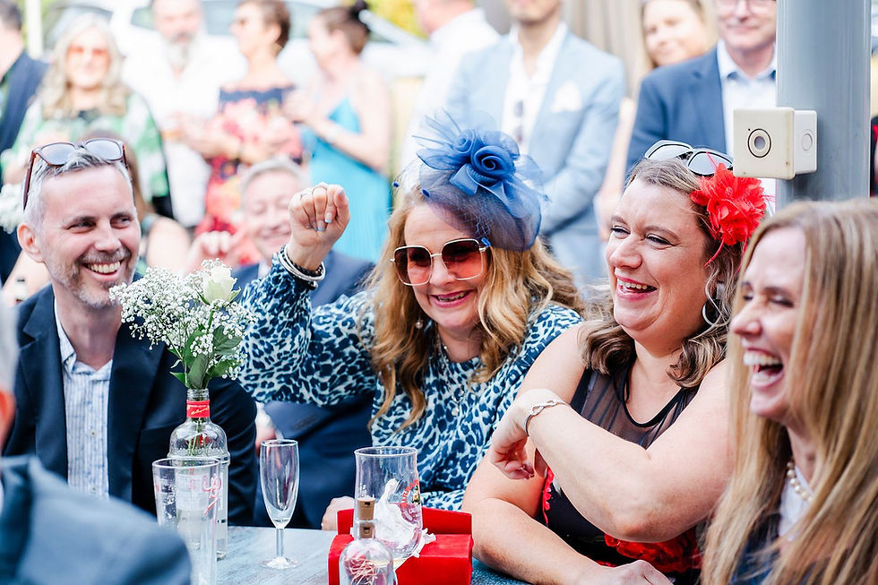 Group of people laughing at a table outdoors. Woman in blue hat raises hand joyfully. Vase with flowers is on the table. Festive atmosphere.