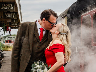 A couple kisses on a vintage train platform, surrounded by steam. She wears a red dress holding flowers; signs read "Ladies Room" and "Booking Office."