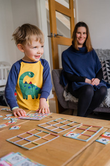 Laughing toddler in dinosaur pyjamas playing with cards on a table while his mum watches from the sofa behind him