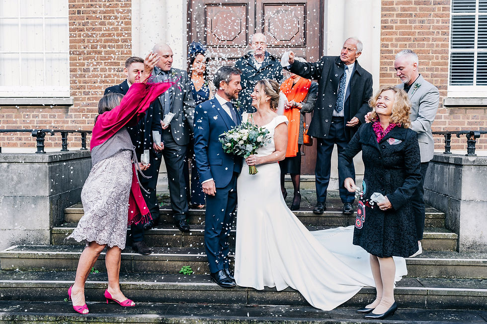 Bride and groom standing on stone steps outside an ornate wooden door, surrounded by guests throwing confetti, at Hertford Register Office