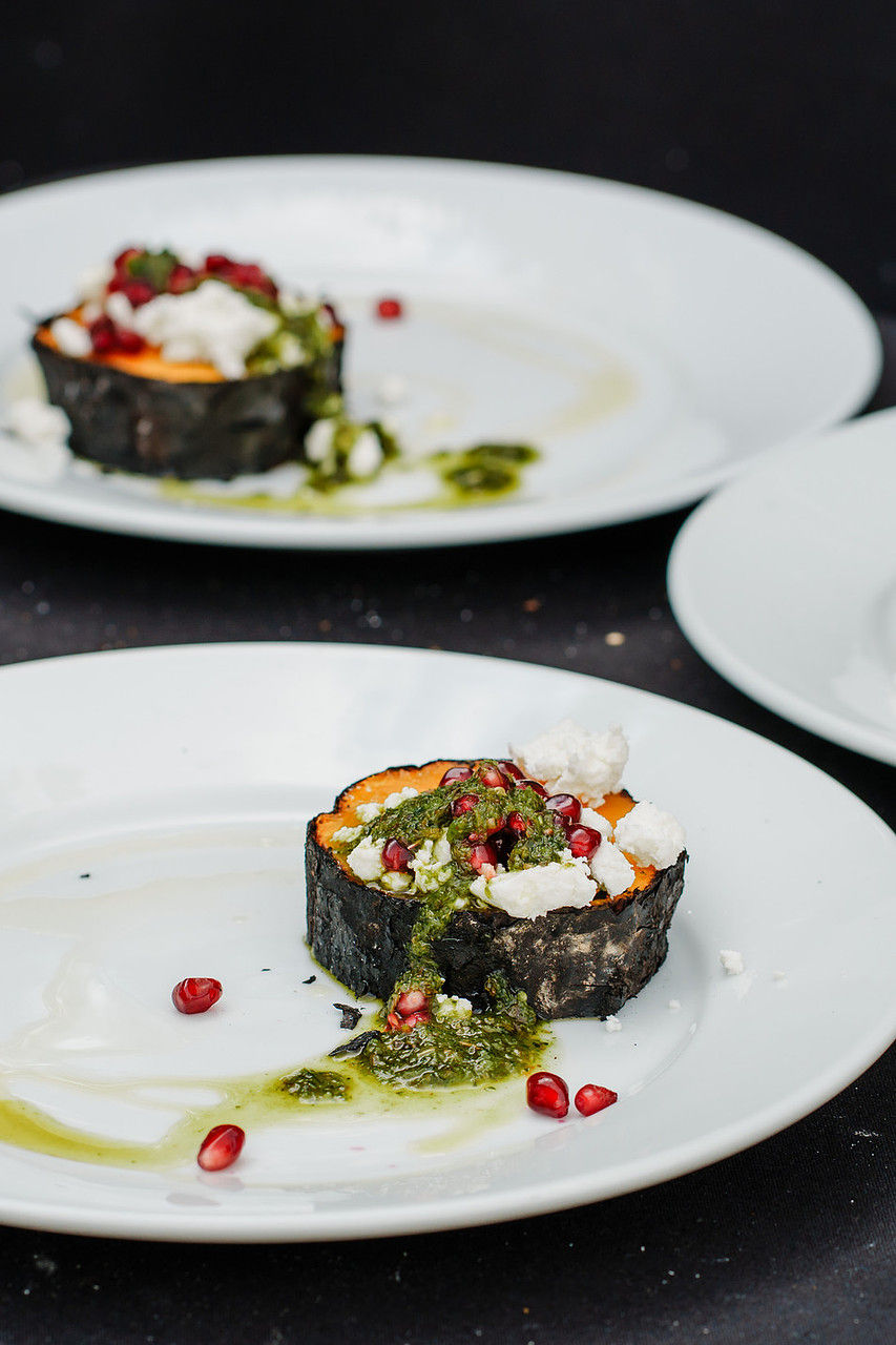 Plated roasted squash rounds topped with pesto, cheese, and pomegranate seeds on white plates against a black background