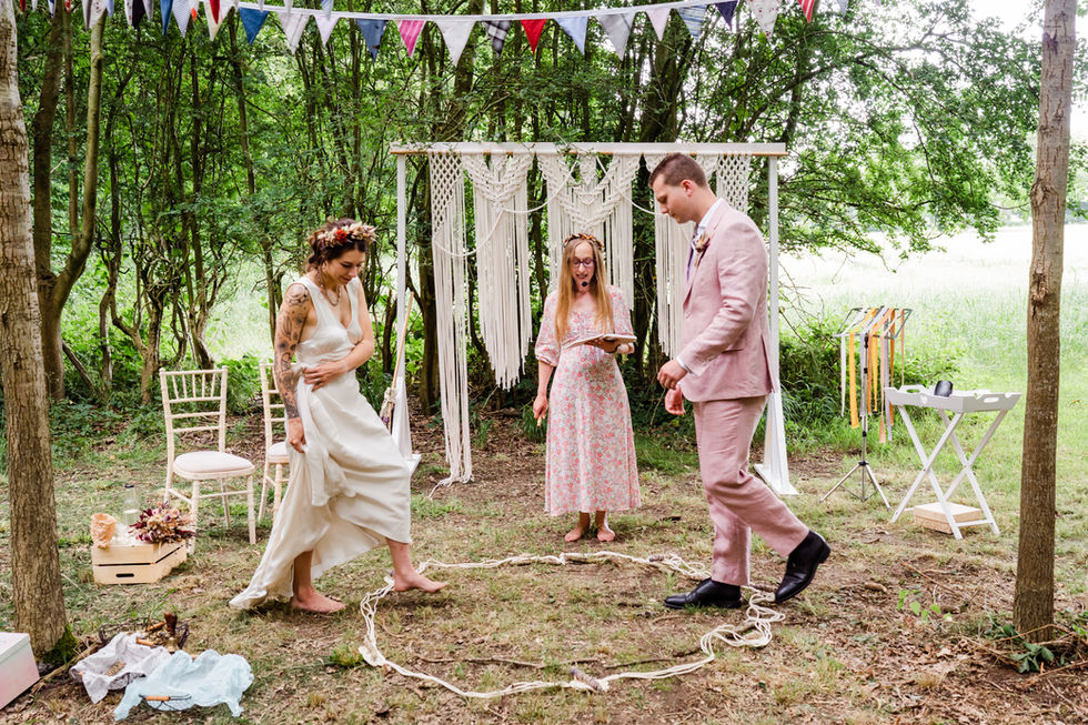 Barefoot bride and groom in a pink suit stepping into a rope circle during their woodland wedding ceremony