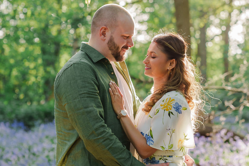 A couple embraces in a sunlit forest. The woman wears a floral dress, the man a green shirt. Blossoms and greenery create a serene mood.