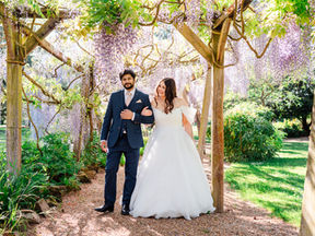 Bride in white gown and groom in blue suit walk under wisteria arbour, surrounded by greenery, on a sunny day, looking happy.
