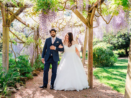 Bride in white gown and groom in blue suit walk under wisteria arbour, surrounded by greenery, on a sunny day, looking happy.