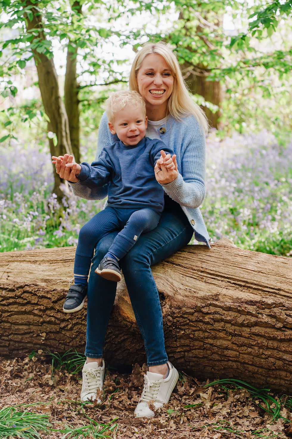 A smiling woman and child in blue sit on a log in a lush, green forest. Both are joyful, surrounded by trees and purple flowers.