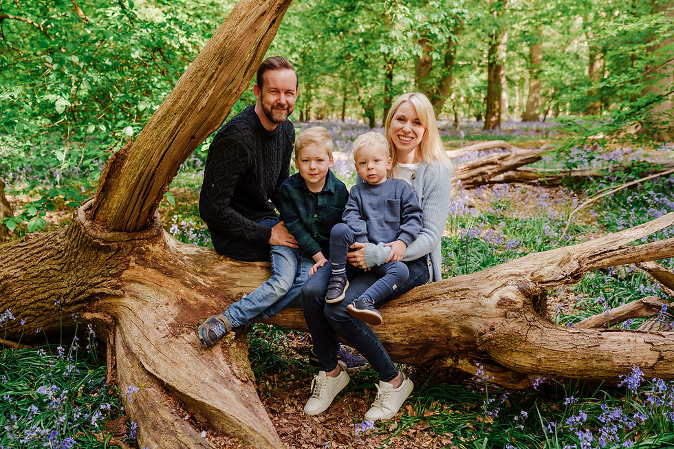 Family of four smiling on a fallen tree in a lush green forest with purple bluebells. Bright, cheerful atmosphere.
