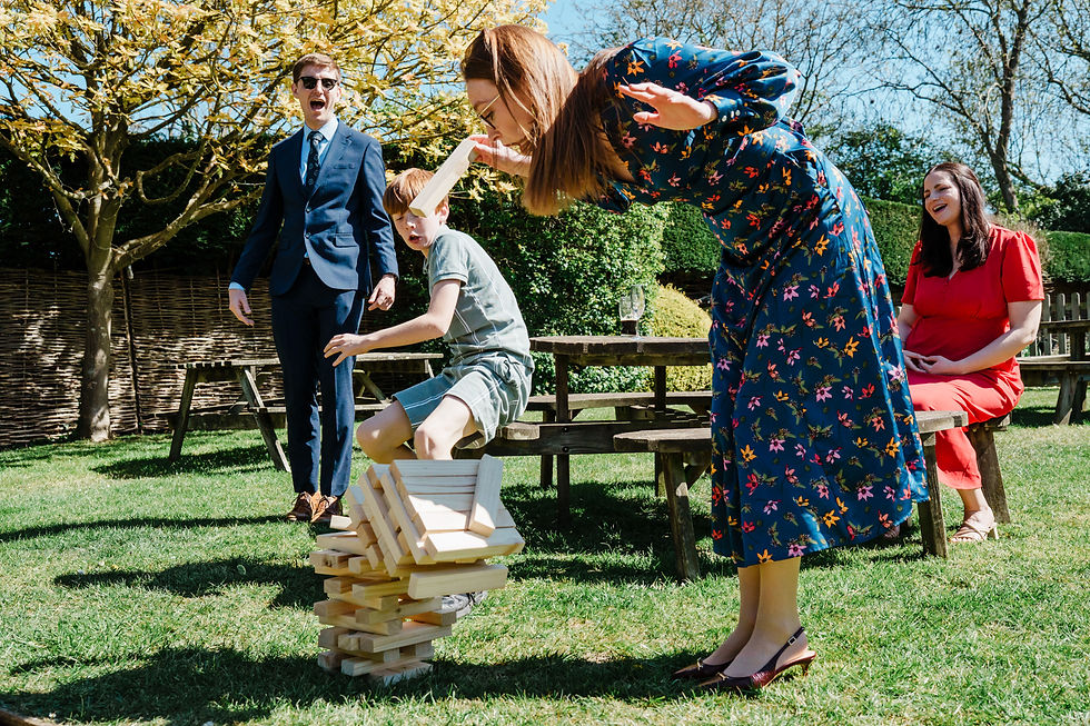 People play giant Jenga on a sunny lawn; a woman in a blue floral dress topples the tower while others laugh.