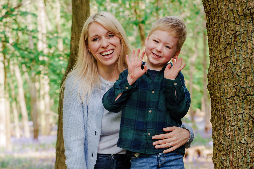 Smiling woman and child in a forest, child raising hands playfully. Bright greenery and tree trunk in the background, projecting joy.