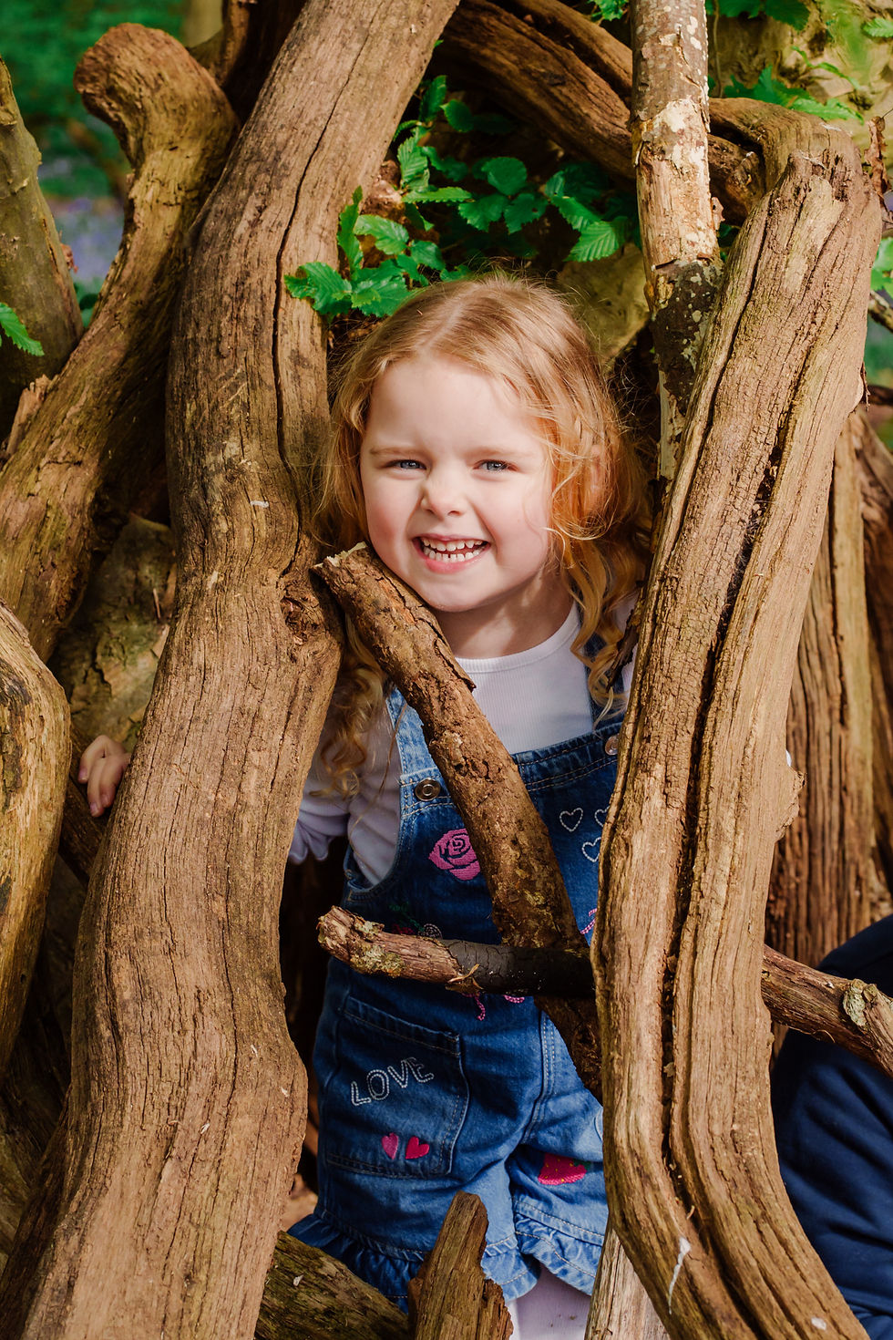 Young girl smiling through twisted tree branches, wearing denim overalls with heart and rose patterns. Lush greenery in the background.