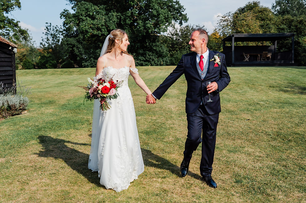 Bride and groom smiling, holding hands in a garden. Bride in white gown with red bouquet, groom in dark suit with red tie. Sunny day.