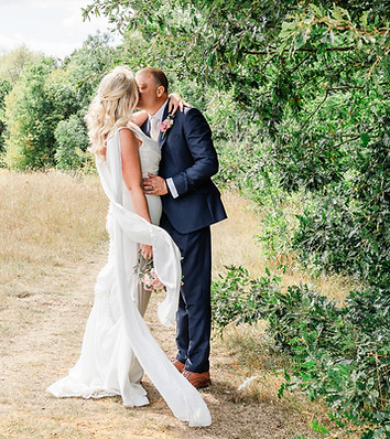 Bride in white gown and groom in navy suit kiss on a grassy path beside trees, holding a b
