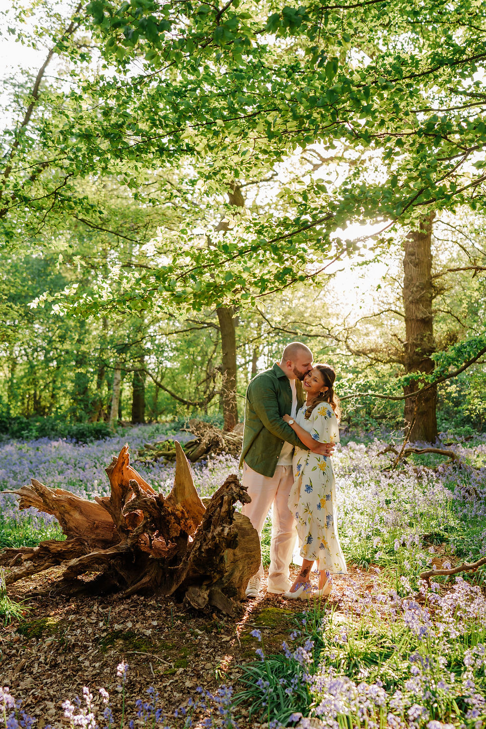 A couple embraces in a sunlit forest with purple flowers and green leaves. The mood is romantic and serene.