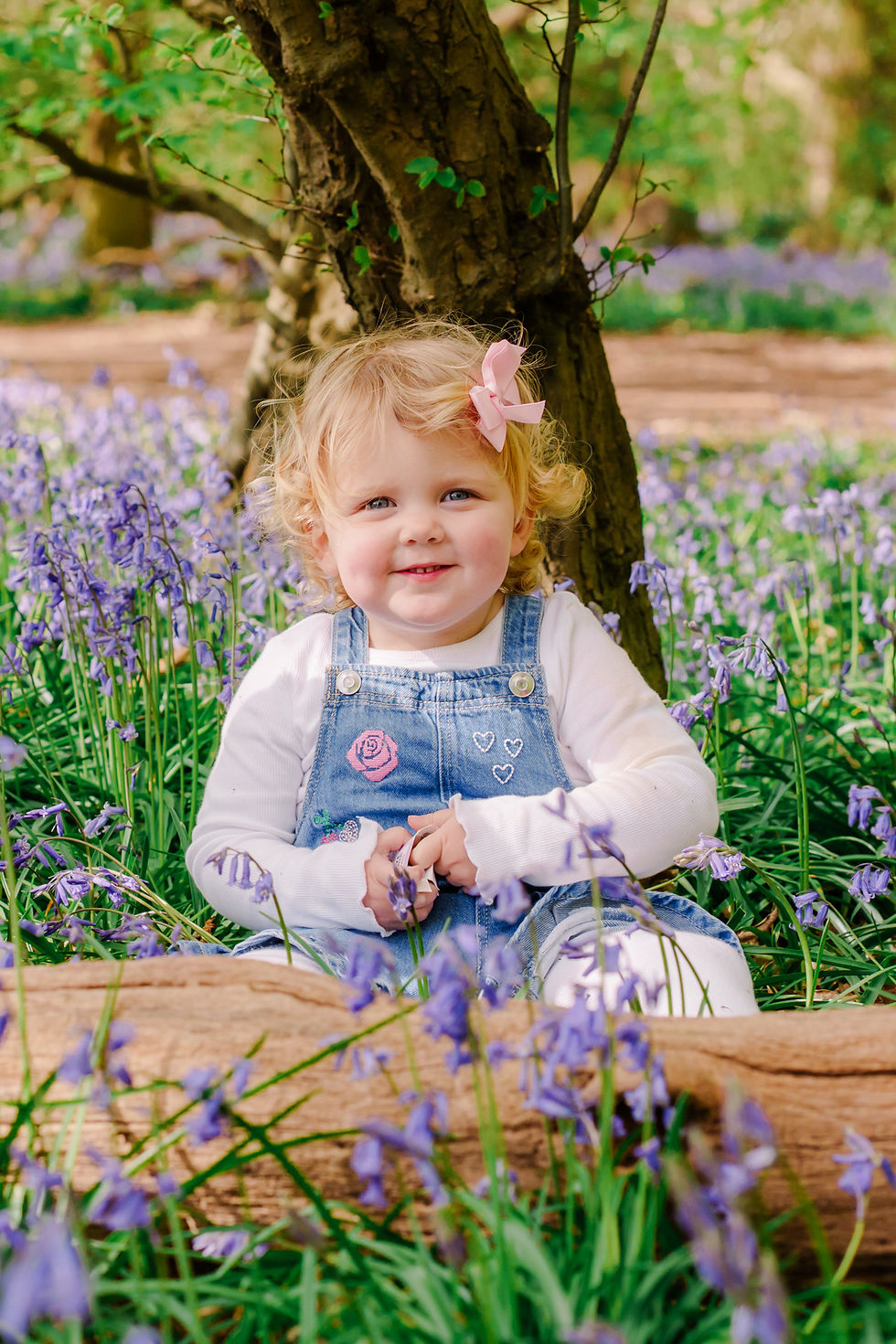 A smiling child with a pink bow sits among purple bluebells under a tree, wearing denim overalls with heart and flower designs.