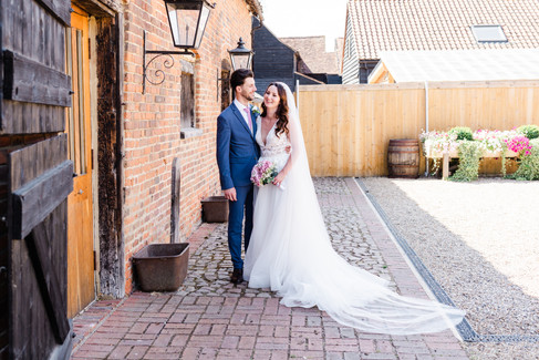 Groom in blue suit standing next to Bride holding pink flowers next to the wall of a barn building