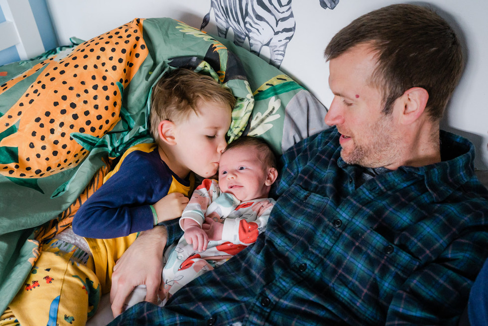 Toddler wearing blue and yellow pyjamas gently kissing a newborn on his temple while they lie together on a bed with their father