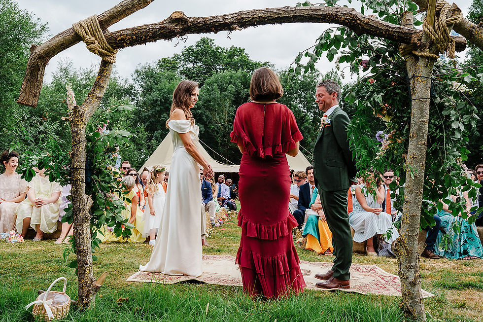 Outdoor wedding ceremony under a rustic arch, with bride in white, groom in green, and officiant in red before seated guests