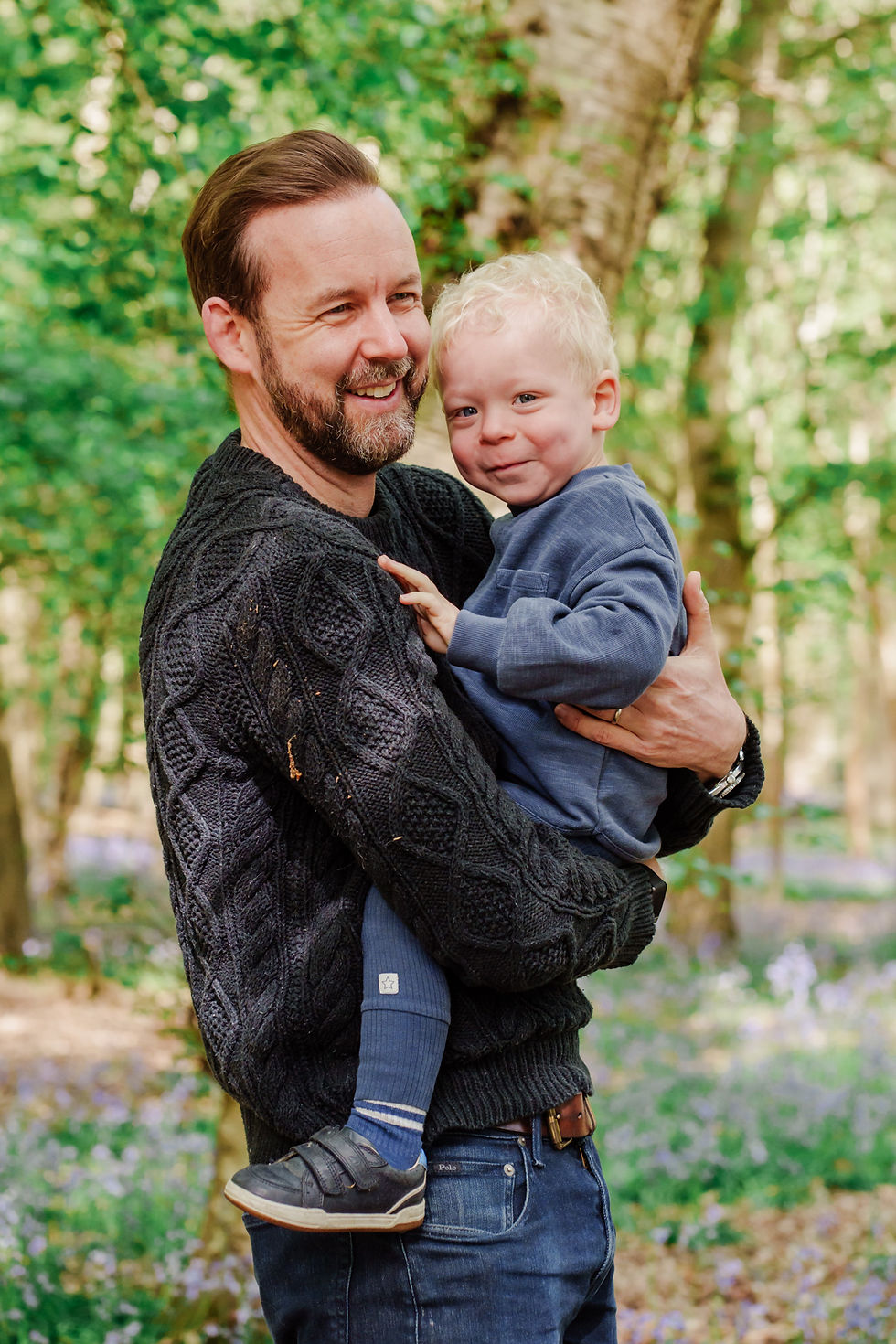 Man in a dark sweater smiling, holding a young boy in blue. They're in a sunlit forest with green leaves and purple flowers. Joyful mood.