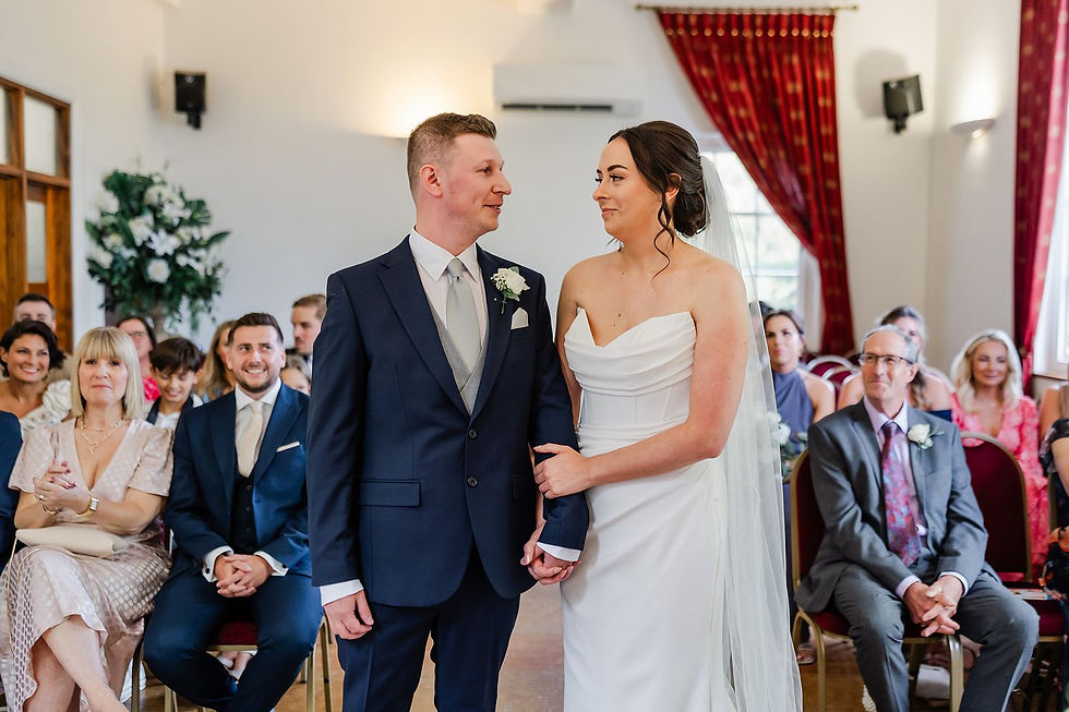 Bride and groom stand together holding hands, smiling at each other. Guests seated in a room with red curtains watch them.