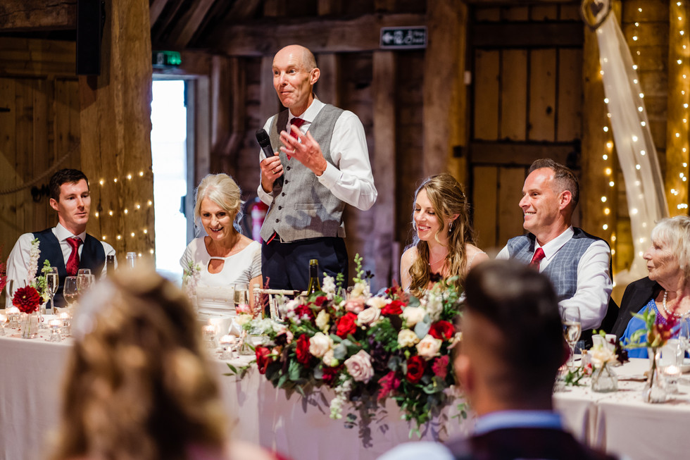 Man in grey waistcoat and red tie standing at the top table in a barn giving a wedding speech